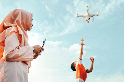 Low angle view of mother flying drone while son playing with it against sky