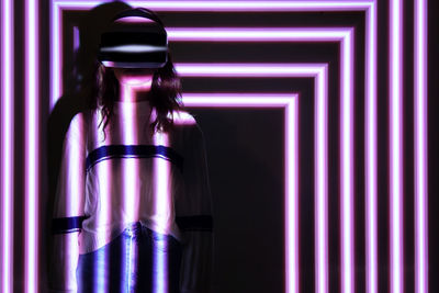 Young woman wearing virtual reality simulator standing against illuminated background