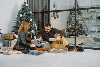 Family with child playing near christmas tree. child unpacking gifts, parents enjoy christmas tree