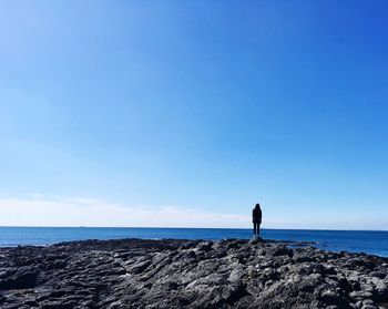 Rear view of woman standing on rock by sea against blue sky