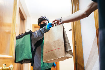 Delivery woman with thermal backpack and face mask giving paper bag with food to crop faceless client through apartment door