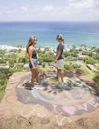 Friends standing on cliff with graffiti near sea against cloudy sky