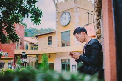 Side view of young man using smart phone while standing by building