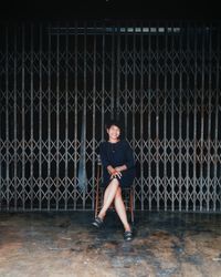 Full length portrait of smiling young woman sitting against closed gate