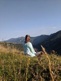 Woman sitting on grass against mountains and sky