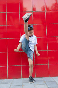 A cute boy in a white shirt raises his leg against a red wall, the football