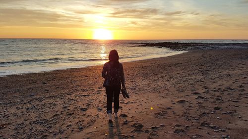 Full length rear view of woman standing at beach against sky during sunset