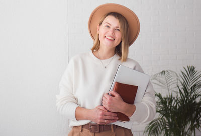 Trendy woman freelancer wearing brown hat holding notebook, smiling and looking at camera