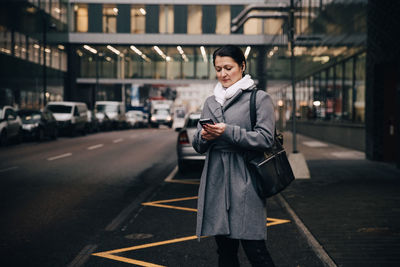 Businesswoman using smart phone while standing on city street against building