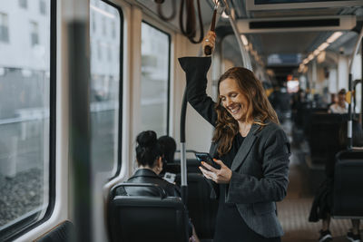 Cheerful female passenger using smart phone while standing in train