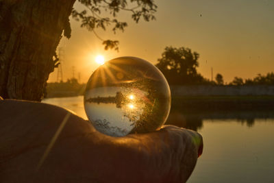 Reflection of person hand holding crystal ball on lake against sky during sunset