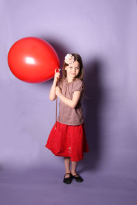 Portrait of smiling girl holding red balloon while standing against purple background