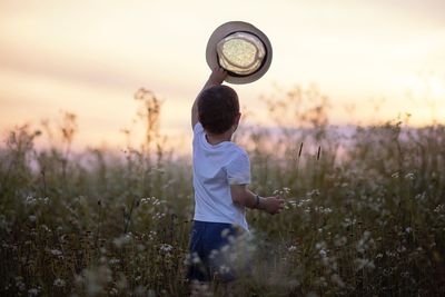 Rear view of boy holding hat while standing on field against sky during sunset