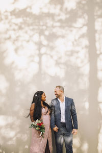 Happy multiracial bride and groom holding hands against wall