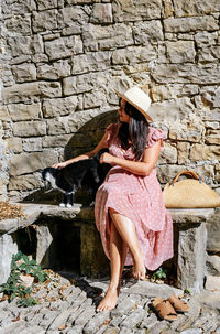 Young woman in pink dress petting a cute cat, stone wall, bench, sunny day, lifestyle. smiling.