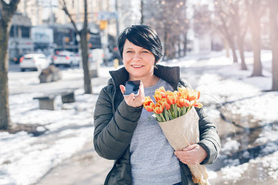 Middle age woman walking on spring street with yellow tulips flowers and talking on audio chat phone