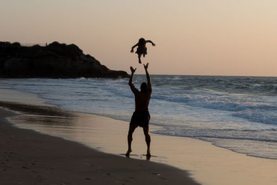 Full length of silhouette man throwing son while standing at beach against clear sky during sunset