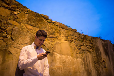 Young man using mobile phone while standing against wall and sky