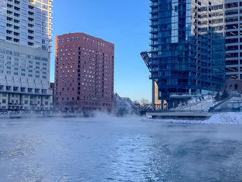 Modern buildings by river against sky in city during winter