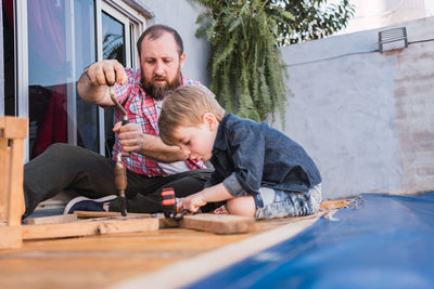 Bearded dad teaching son with hammer working with wood while sitting on boardwalk on weekend