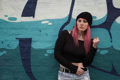 Young tattooed woman with pink hair standing against graffiti wall