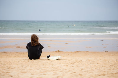 Rear view of man sitting by surfboard at beach