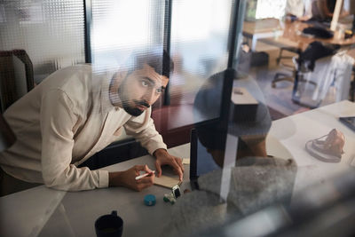 Businessman writing in note pad while discussing with male colleague at desk in creative office