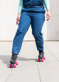 Cropped unrecognizable lady with short hair in denim overall riding roller skates on pavement against building wall