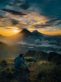 Man sitting on mountain against sky during sunset