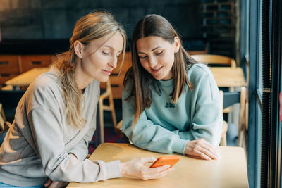 Two modern women surf the internet using a mobile phone while sitting in a coffee shop.