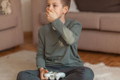 Boy with hand covering mouth holding video game remote control in living room at home 