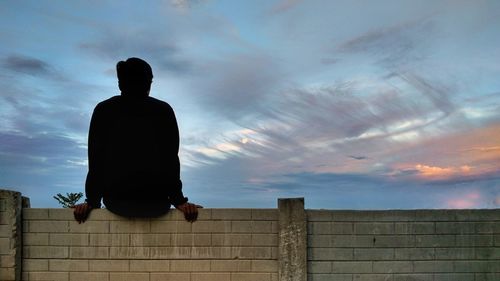 Rear view of man sitting on wall against sky