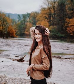 Portrait of smiling young woman with long hair standing against lake in forest