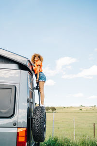 Full body black female tourist with curly hair looking up and climbing up ladder on modern rv against blue sky and green field in countryside