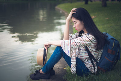 Side view of worried woman with backpack sitting by lake in park