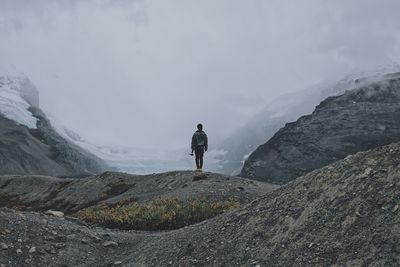 Rear view of man standing amidst mountains against sky during foggy weather