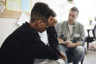 Sad teenage boy sitting by male counselor discussing in school office