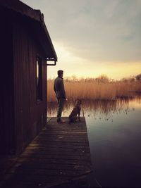 Man with dog standing on deck of stilt house by lake during sunset