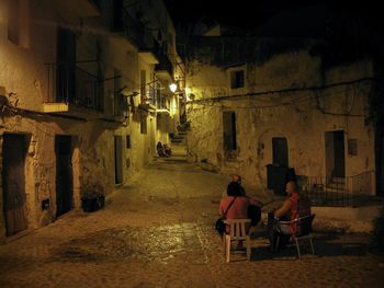 Rear view of people sitting on street against buildings at night