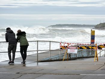 Rear view of man and woman standing by fence and sea against sky