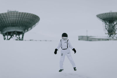 Unrecognizable cosmonaut in spacesuit standing in snowy valley in winter on background of huge radar antennas in svalbard