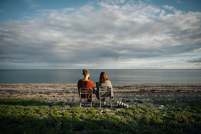 Rear view of woman sitting on beach looking at sea against sky