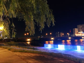 Illuminated street by buildings against sky at night