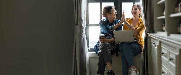 Young woman using mobile phone while sitting on window