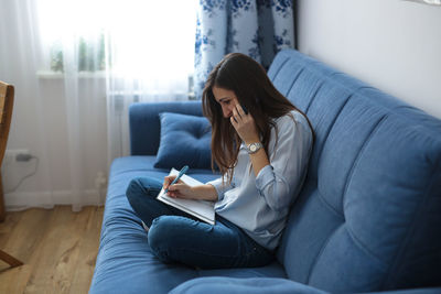 Beautiful woman talking on the phone and writing in a notebook sitting on a blue sofa in the room