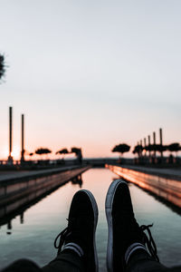 Low section of person by river against sky during sunset