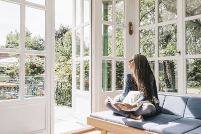 Woman sitting with book on lounge in winter garden looking through opened terrace door
