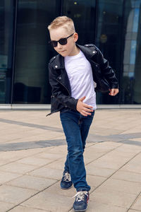 Baby boy in sunglasses and black leather jacket smiling on the background of the glass building