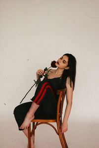 Sensuous woman holding rose while sitting on chair against white wall