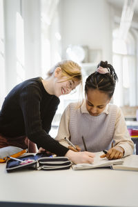 Smiling girl with female friend writing in book while studying in classroom